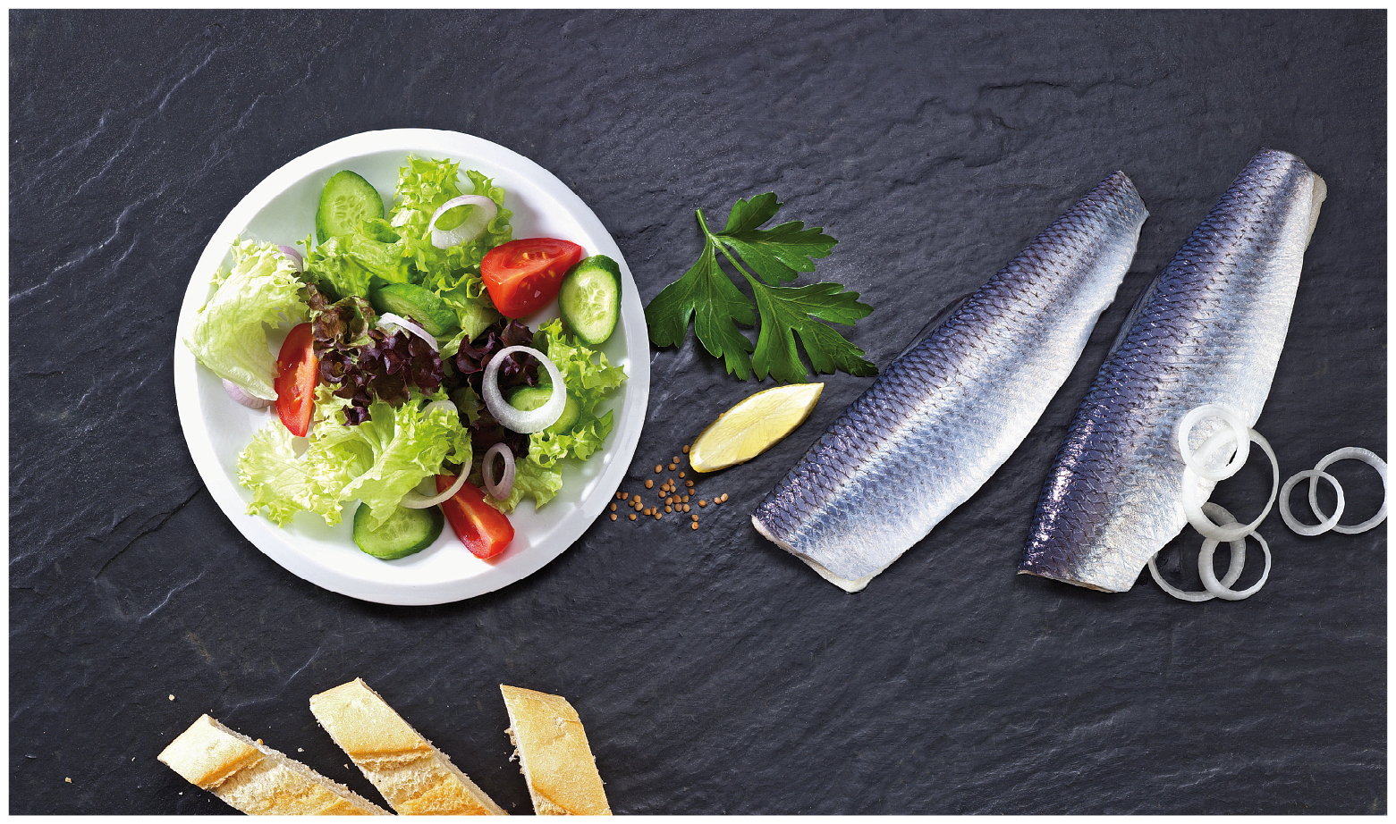 Frischer Hering mit Salat und Baguette auf Schieferplatte Zwei Heringfilets mit Zwiebelringen, gemischter Salat mit Tomaten und Gurken, Baguettescheiben und Zitrone auf dunklem Hintergrund